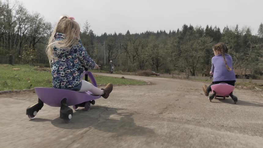 Two little girls riding down a hill on scooters on a trail at the park