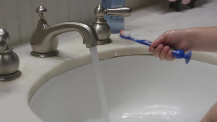 Blue toothbrush being rinsed under running water from faucet in bathroom