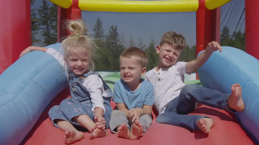 Three white children smile while sitting on an inflatable play area