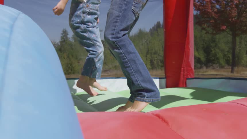 An adult and child both wearing blue denim and white are both barefoot and jumping in the blue red and green bouncy house