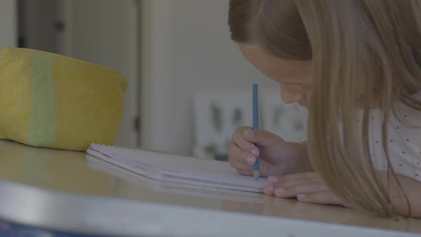 A young girl uses a blue color pencil to write in a notebook