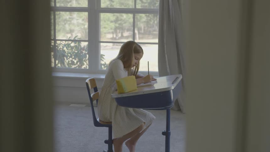 A young girls sits at a chair fixed to a desk in a bedroom and is drawing with a pencil in a notebook