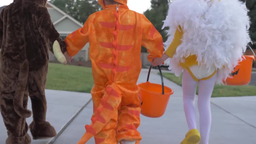 Three children in fancy dress outfits walk down a street to trick or treat on halloween