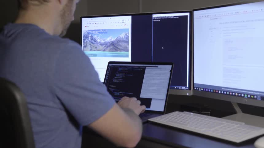 A man types on a laptop in front of two large monitors