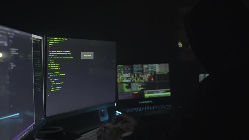 A man sits in a dark room working on a computer with three monitors