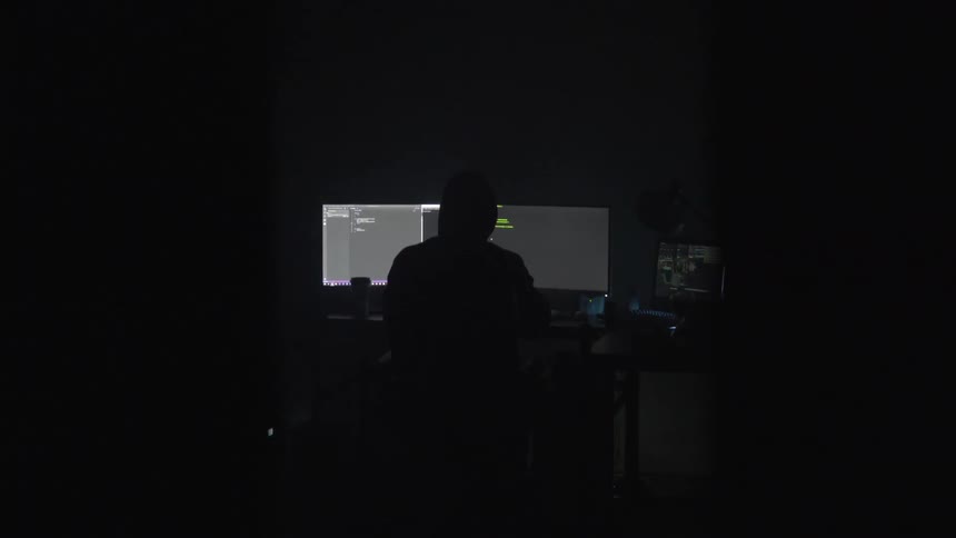 A silhouetted person sits at a desk with multiple computer monitors in a dark room