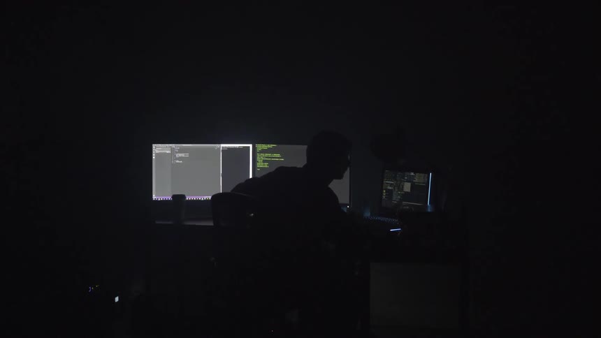 I man sits in a dark room at a desk looking at three computer monitors