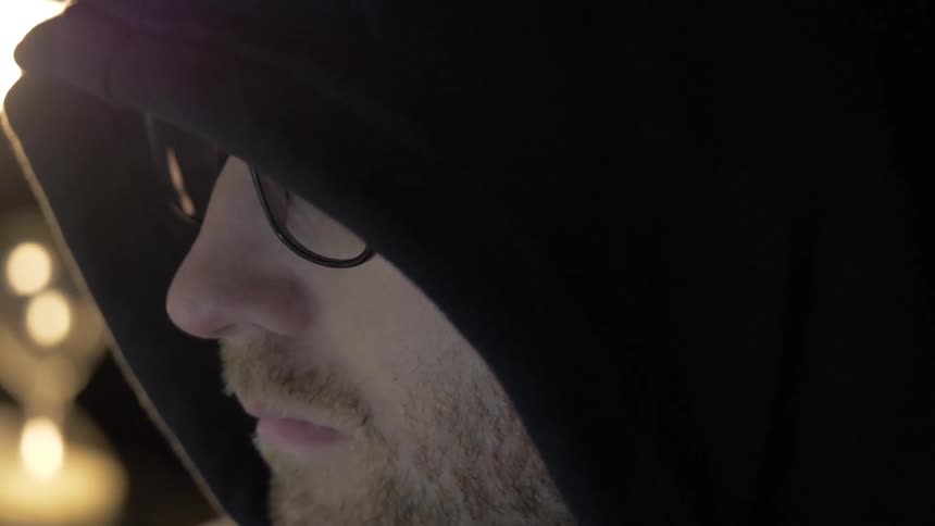 A man wears a black hoodie pulled over his head while looking at a screen