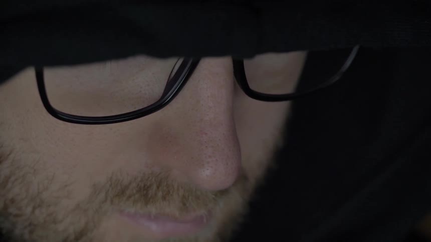 A man wearing glasses and a low slung hat works on something below screen