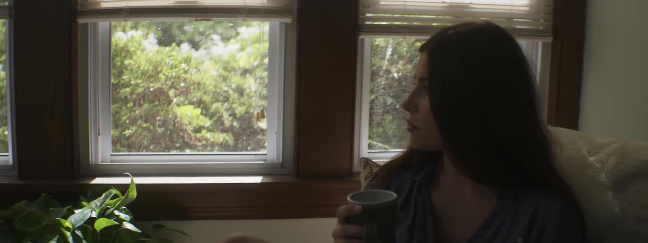 A brunette woman sitting down with a book and coffee cup turns to look out the window