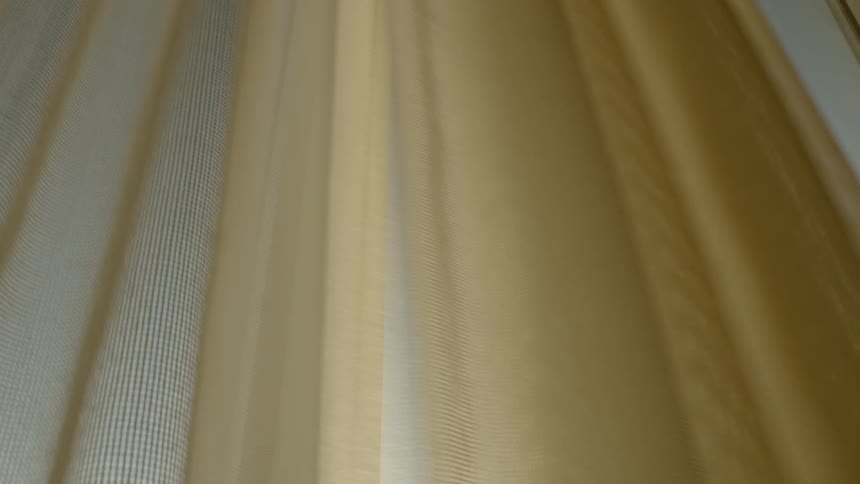 A long beige fabric curtain is pulled aside to reveal a window behind it and a clear blue sky outside