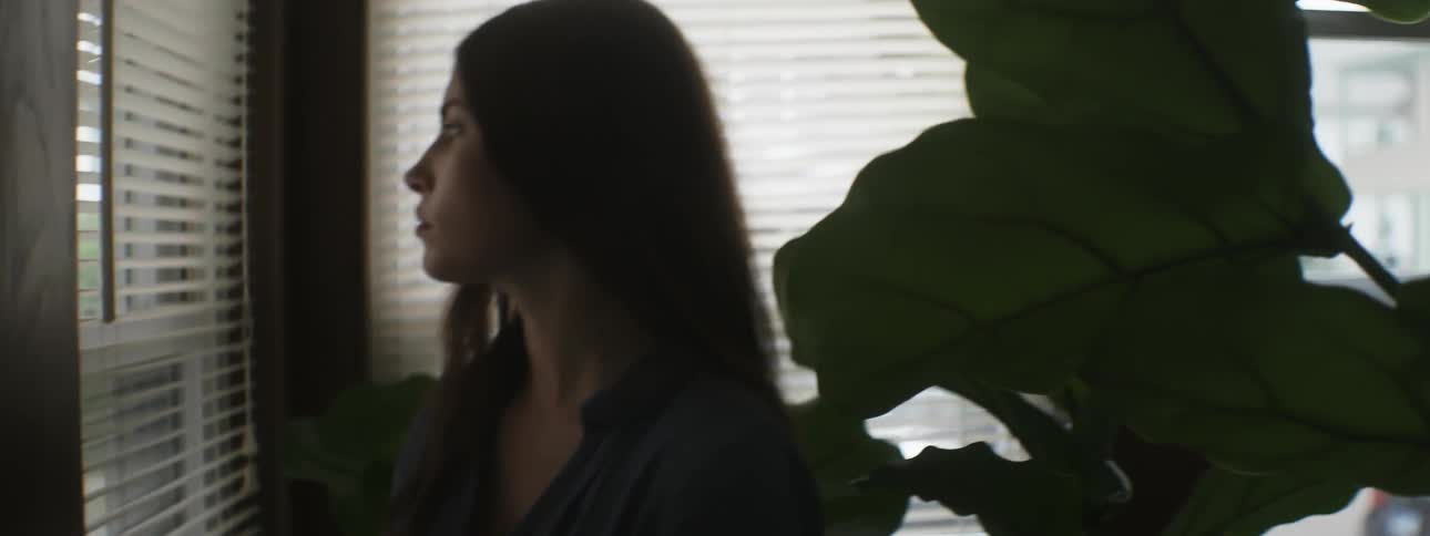 A young woman opens the blinds of window and stares out at the neighborhood outside