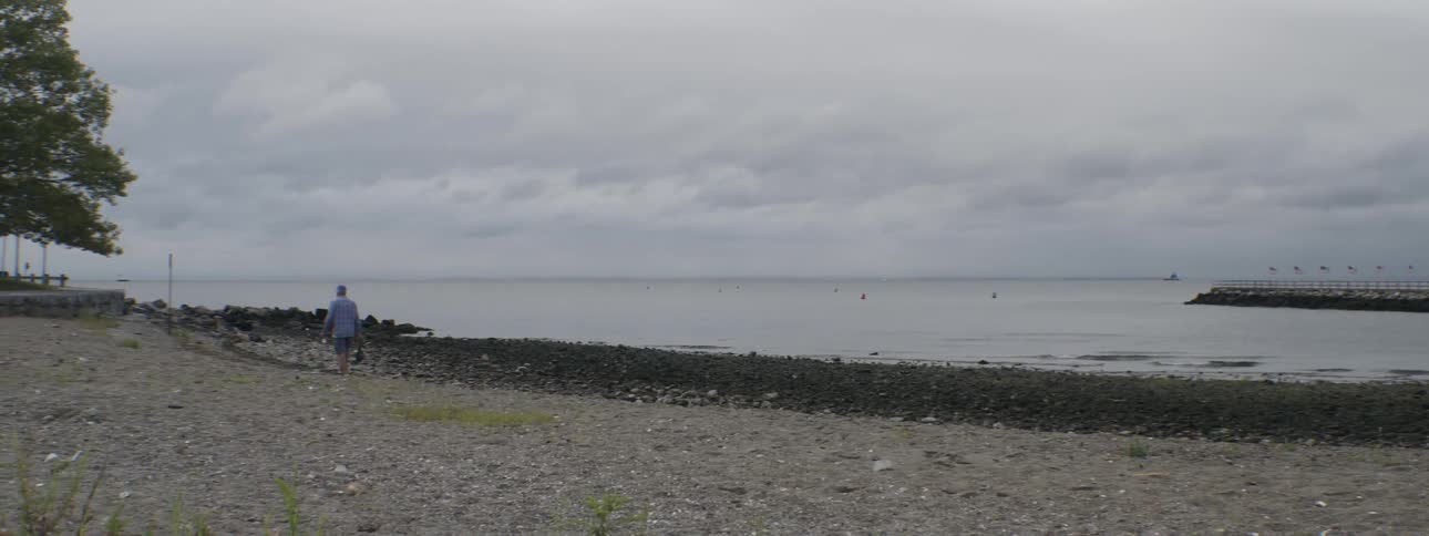 Man holding a fishing pole walking down a stony beach on a cloudy day