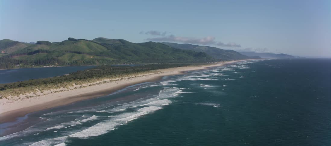 Camera flies along an ocean coast covered by sand with mountains in the background