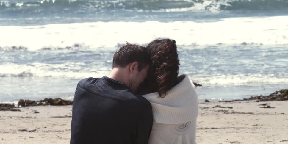 A man burying his face in a woman's hair as they sit on a beach watching waves roll in on a chilly day