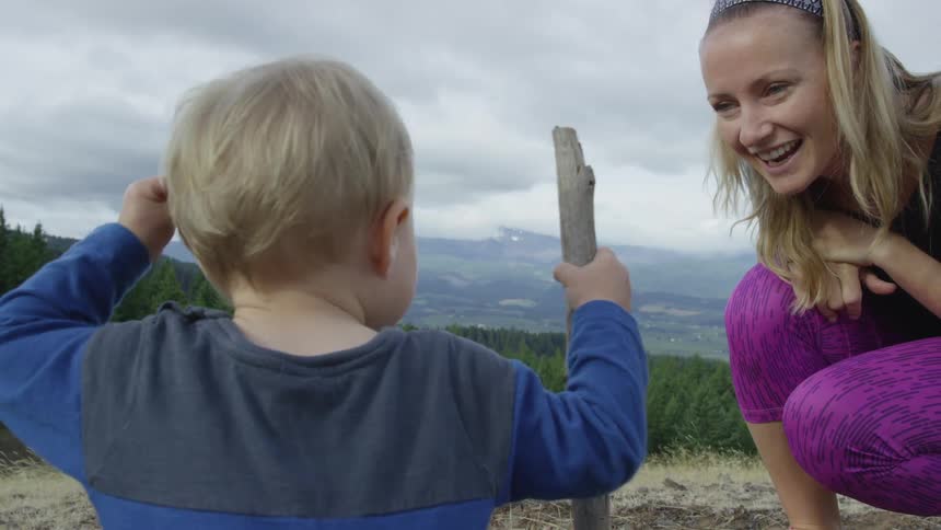 A toddler and his mother play outdoors in an area with forest trees and mountains