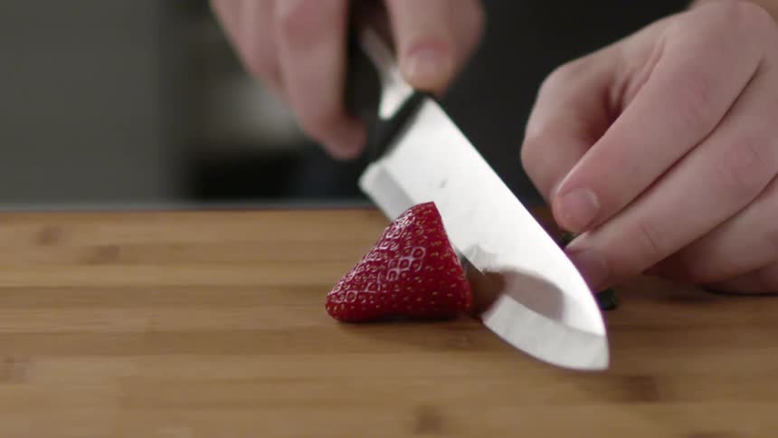 The hands of a person using a knife to cut the stem and top off of a strawberry