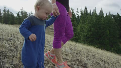 A young mother holds her toddler's hand while they explore the woods while walking around on a nature hike