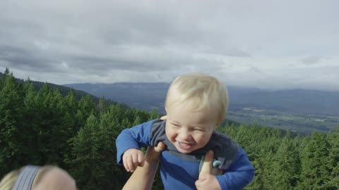 A blonde woman lifting a smiling blonde toddler with giant pine trees, mountains and a cloudy sky in the background