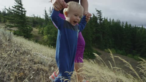 A young mother helps her toddler to walk down a grassy hill in the wilderness on a hike