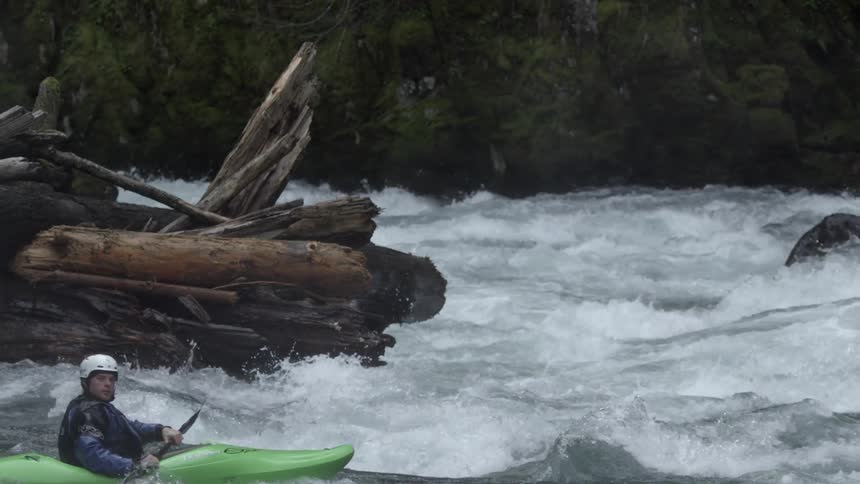 Slow motion shot of a whitewater kayaker paddling past a log jam at the corner of a large rapid