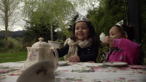 Two little girls with tiaras are having a tea party on a table on the lawn