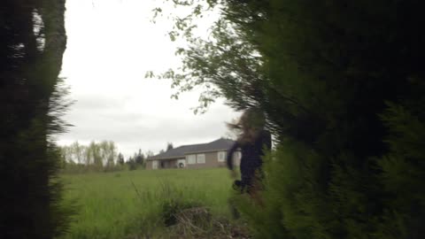 Two girls are running near some evergreen trees at the edge of a large grassy field