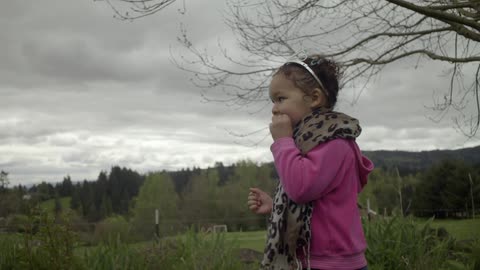 A little girl stands by a tree and looks around at the surrounding trees and hills on an overcast day