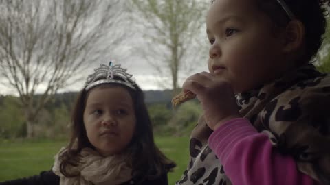 A little girl eats a snack while another girl wearing a crown watches her