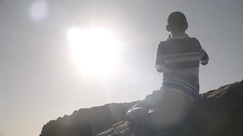 A boy sitting on some big rocks takes pictures while the sun glares down on him