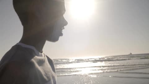 A young boy walking along a beach as waves come on shore and the sun glares in the background