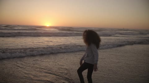 A young girl is standing in the water at sunset as the small waves chase after her