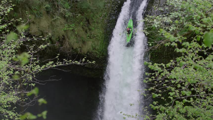 Slow motion shot of a man in a green whitewater kayak dropping a massive 100 foot waterfall in a green verdant river canyon