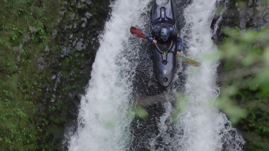 Tight slow motion shot of a man in a black kayak dropping a huge waterfall deep in a lush green canyon