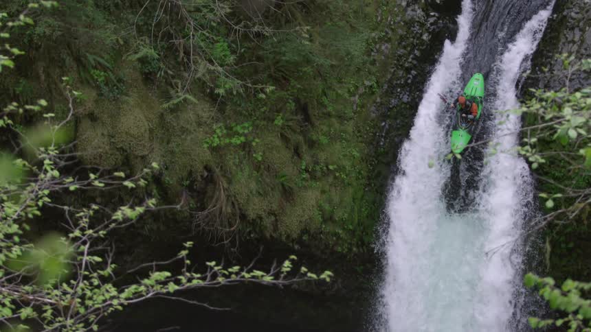 Slow motion shot of a whitewater kayaker dropping a hundred foot waterfall deep in a lush green canyon