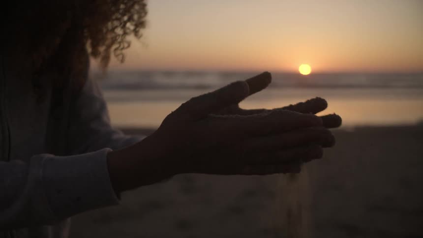 A woman on the beach runs sand through her fingers at sunset