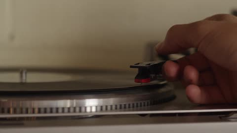 A hand raises the arm of a record player and places the needle at the start of the record