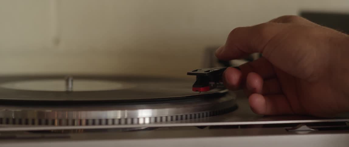 A hand raises the arm of a record player and places the needle at the start of the record