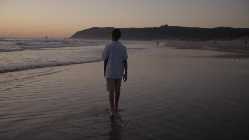 A boy walks along the beach in the water at dusk, as the little waves roll in at his feet