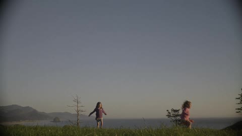 Three children are running and jumping in a meadow above a lake