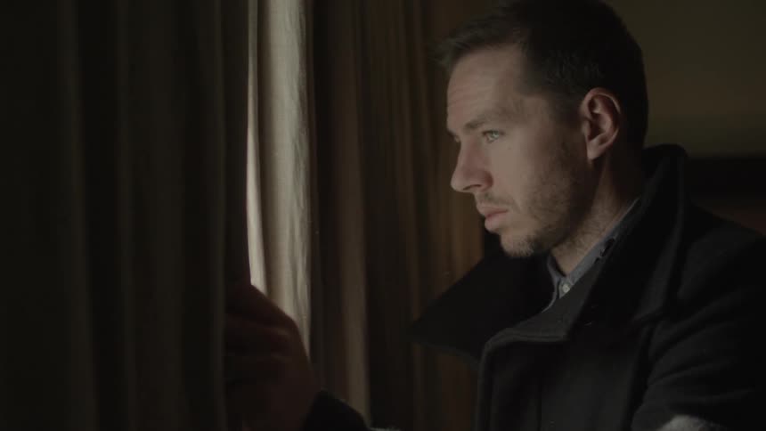 A man in a pea coat approaches a curtain in a room, looks out and closes his eyes