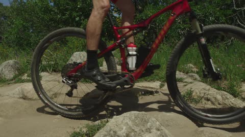 A person rides their bicycle over rocks and sand near green grass and bushes