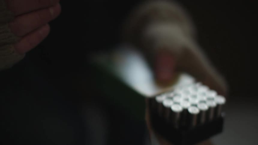 Hands in fingerless gloves remove a single bullet from a box, and after inspection, returns the bullet to the box