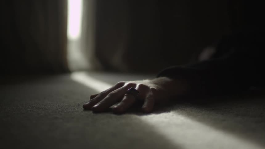 A person's hand lies motionless on the carpeted floor as a shard of light shines in through the curtains