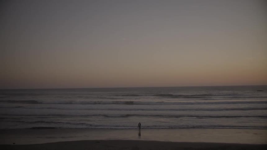 A person standing at the water's edge on a beach during sunset as the waves roll to shore