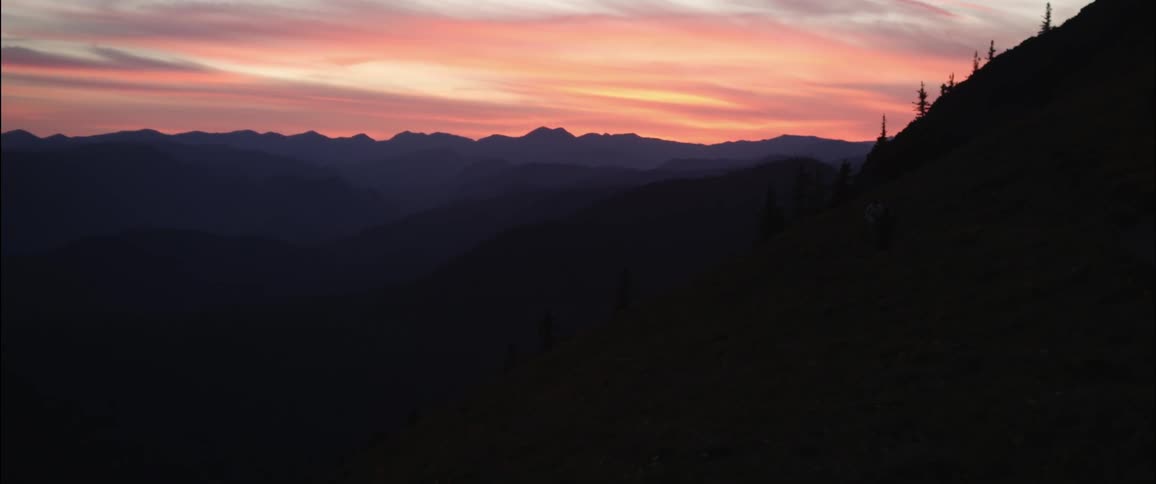 The sun setting behind mountains as people walk along a steep slope