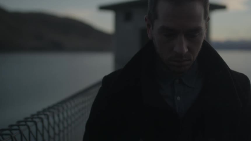 A man in a pee coat walks away from a small building that sits out on the water