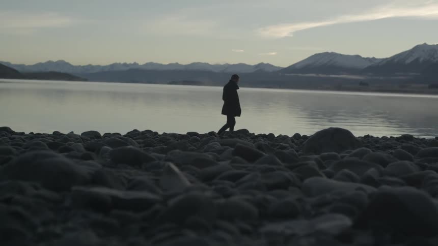 A man walking along a rocky shoreline with mountains and a body of water in the background