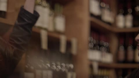 A young man and woman talking in front of a wine display when he hands her a bottle of wine and they give each other a high five