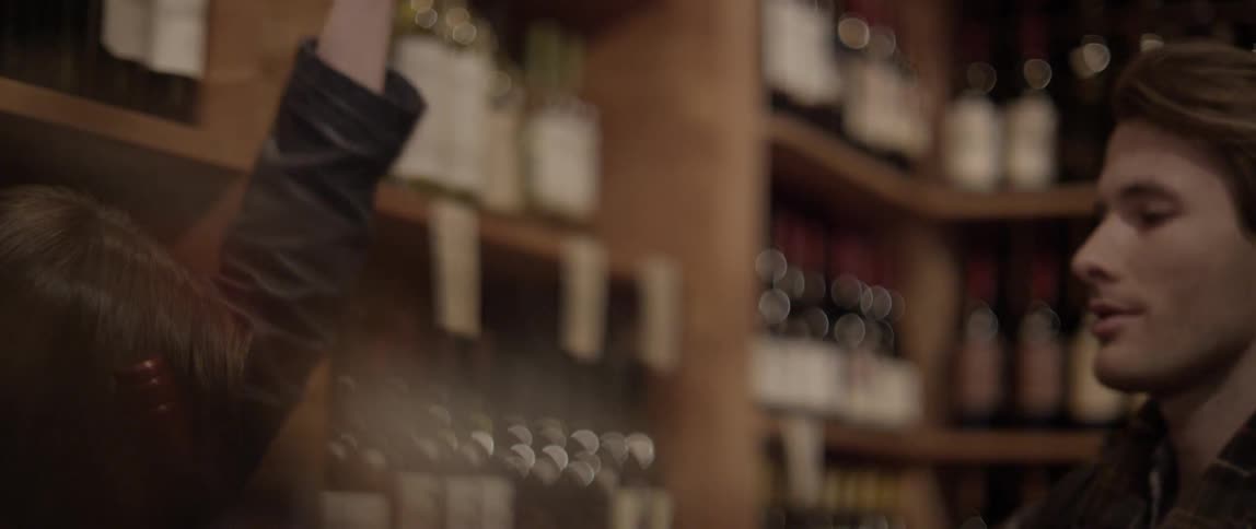 A young man and woman talking in front of a wine display when he hands her a bottle of wine and they give each other a high five